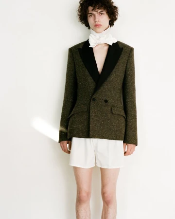 Young male model with dark curly hair wearing an olive green blazer with black lapels and white shorts against a white background