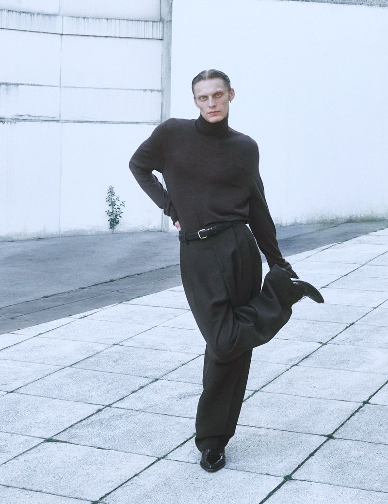Man in turtleneck sweater and wide-leg pants poses with leg raised against a white wall on a concrete surface in an outdoor setting.