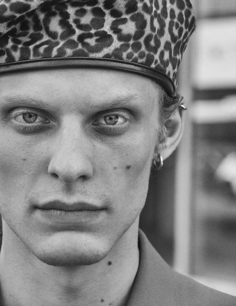 Portrait of an adult male with striking eyes, wearing a leopard-print hat and a tailored jacket, captured in a monochrome photo studio shot.