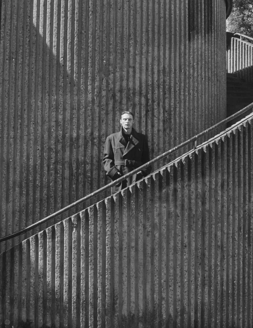 Monochrome portrait of a man in a trench coat standing on concrete stairs in front of a fluted concrete building facade with shadows.