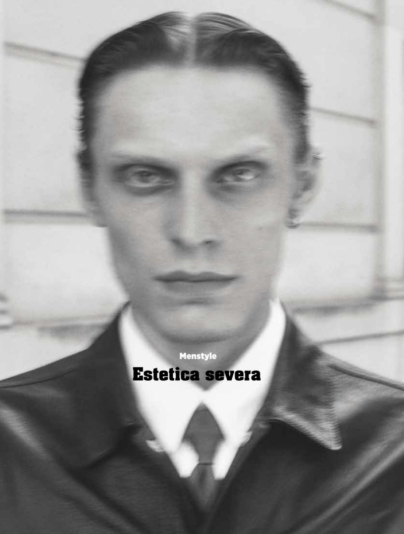 Monochrome close-up of a serious adult male with slicked-back hair wearing a dark jacket, white shirt, and tie in a studio shot.