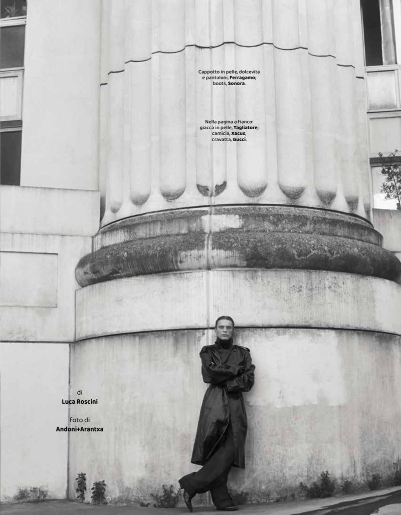 Monochrome fashion portrait of a slender adult leaning against a fluted column, wearing a long coat and wide-legged pants outdoors.