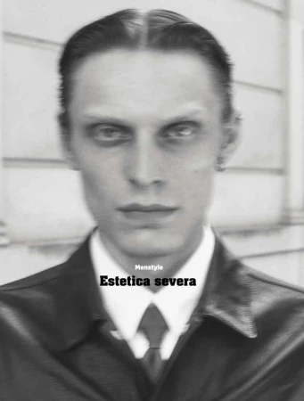 Monochrome portrait of a man in a dark jacket, white shirt, and tie against a building exterior wall.