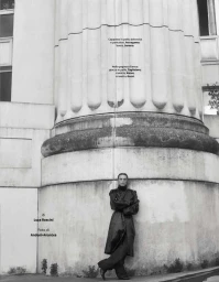 Model wearing a dark trench coat leaning against a large neoclassical architectural column for an editorial shoot.