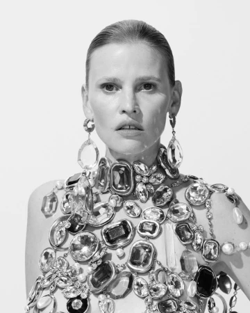 Close-up monochrome portrait of a model wearing an elaborate jeweled collar and statement crystal earrings against a plain background