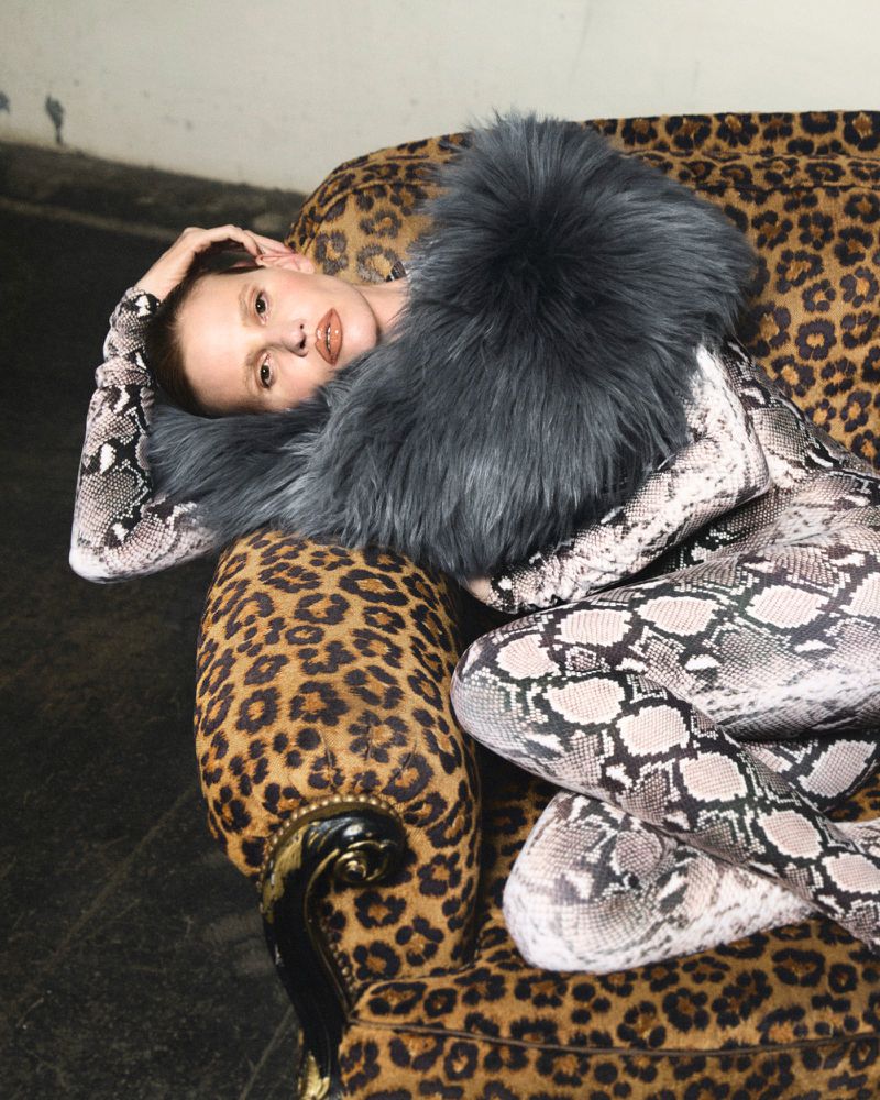 Adult female in snake-print body suit reclines on leopard-print couch with gray fur collar. Studio shot with textured background.