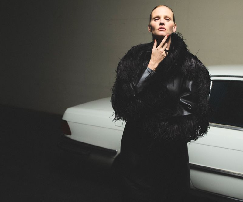 Elegant woman in feather-trimmed jacket poses by vintage white car in moody studio setting. Dark fashion shot with classic automobile.