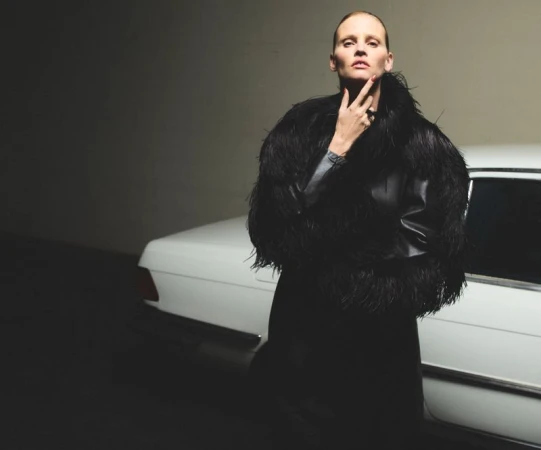Elegant woman in feather-trimmed jacket poses by vintage white car in moody studio setting. Dark fashion shot with classic automobile.