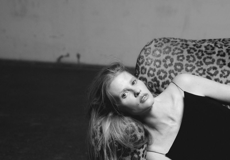 Black-and-white portrait of a woman lying on a leopard-print surface, wearing a black top, with a neutral expression and long hair.