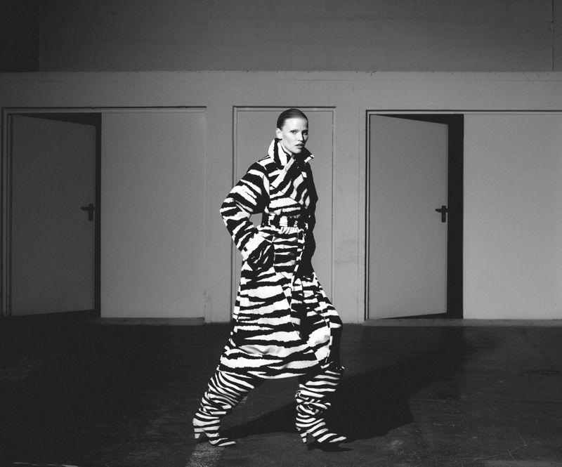 A woman in a zebra-striped outfit poses in a wide shot against a backdrop of closed white doors in a sparse, neutral-toned interior space.