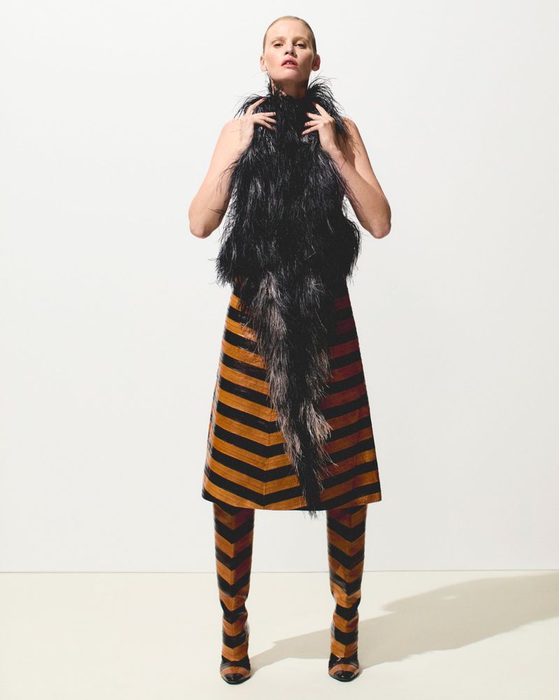 A woman stands in a studio wearing a black feather top, striped skirt, and matching striped boots against a plain white background.