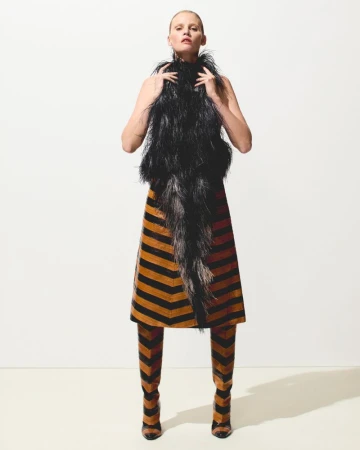 A woman stands in a studio wearing a black feather top, striped skirt, and matching striped boots against a plain white background.