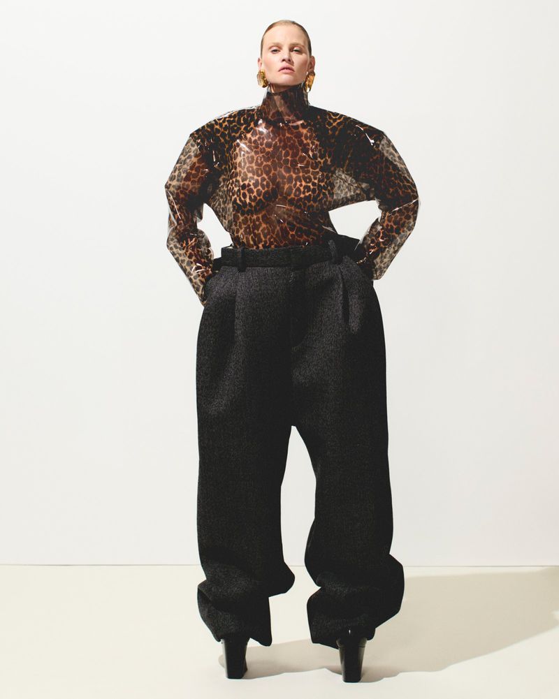 A blonde woman in a leopard-print top and oversized trousers stands confidently against a white backdrop in a fashion-forward pose.