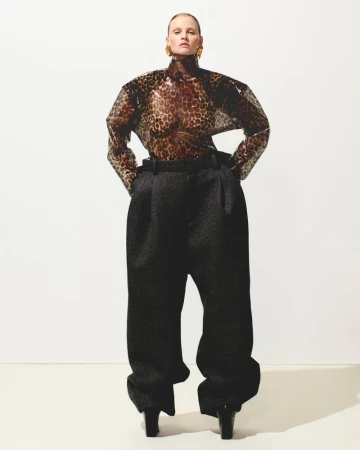 A blonde woman in a leopard-print top and oversized trousers stands confidently against a white backdrop in a fashion-forward pose.