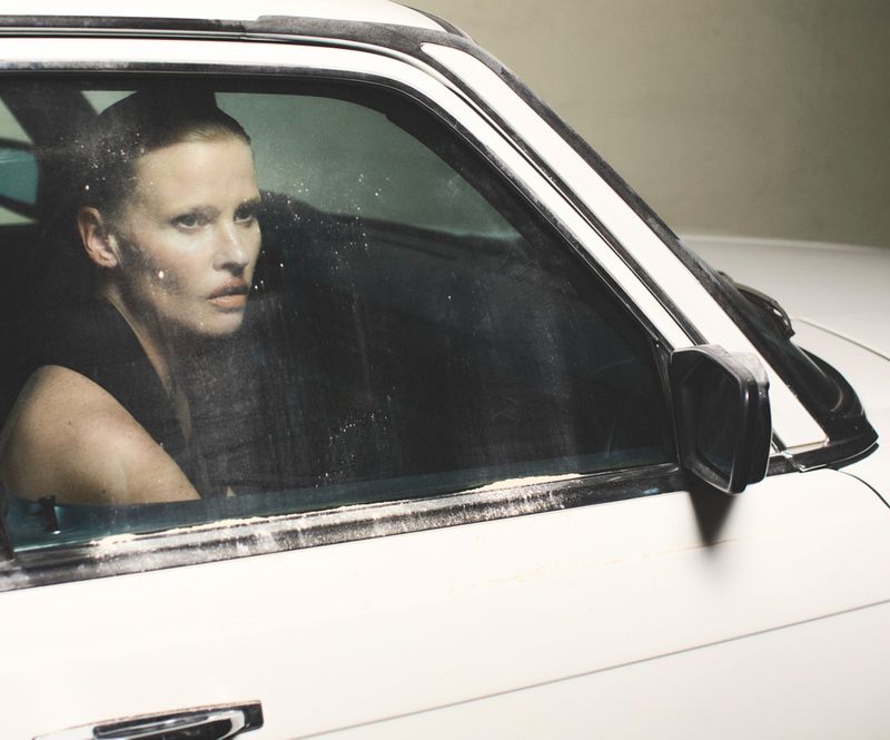 A woman with short hair is seen through the car window. The photo captures the model's stark gaze, creating a dramatic effect.