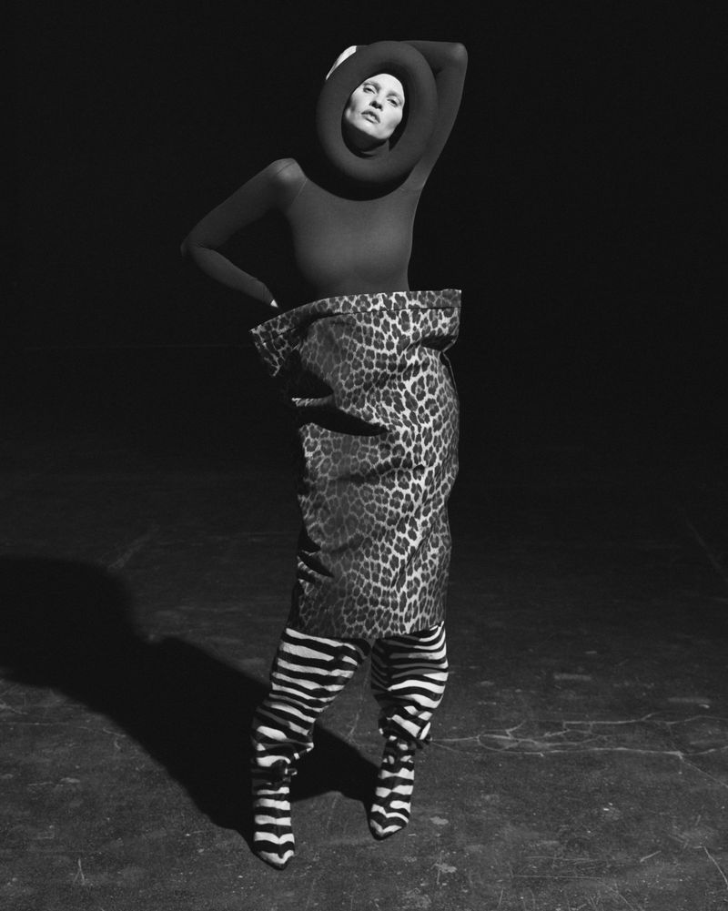 Monochrome portrait of a woman in a black turtleneck, zebra-striped leggings and boots, and a leopard-print skirt in a dark studio.
