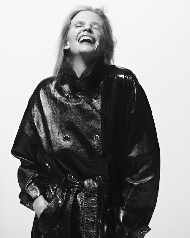 Black and white portrait of a smiling adult woman in a shiny black jacket with a belt, posing against a bright white backdrop.