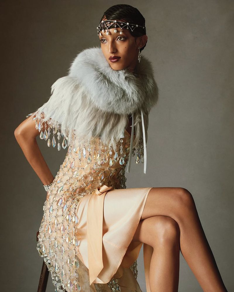 A woman with a jeweled headpiece and feathered collar is sitting in a flapper-style dress adorned with decorative crystals against a grey backdrop.