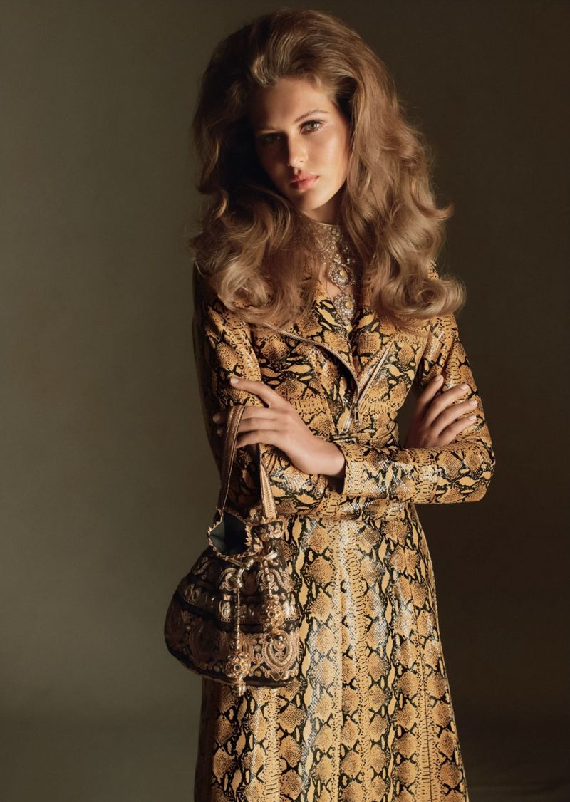 Portrait of a blonde woman in a snakeskin-print coat and carrying a purse, standing in front of a blurred neutral background.