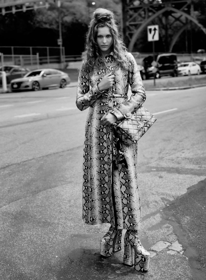 Black and white outdoor shot of a slender woman wearing a snake-patterned coat and platform shoes, holding a matching purse.