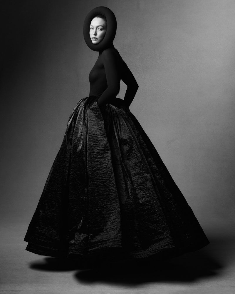 Elegant studio portrait of a woman in a black high-necked top and voluminous skirt, posing against a dark backdrop with an avant-garde headpiece.