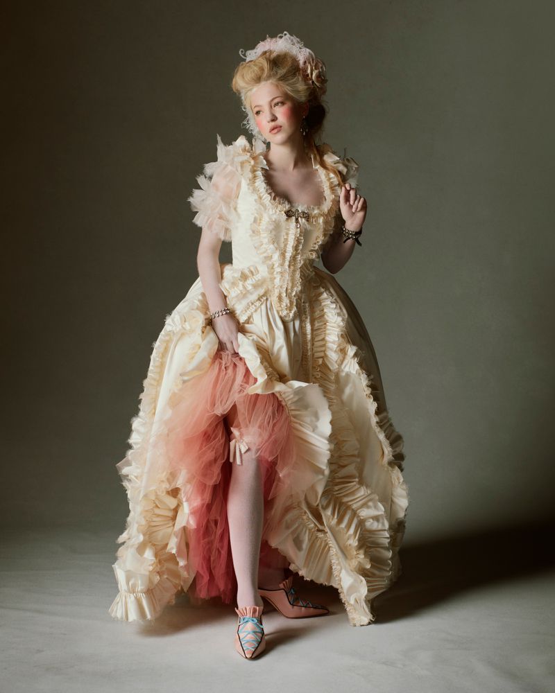 Elegant woman in a ruffled vintage-style gown with lace details poses in a studio. She is holding up the edge of the dress.