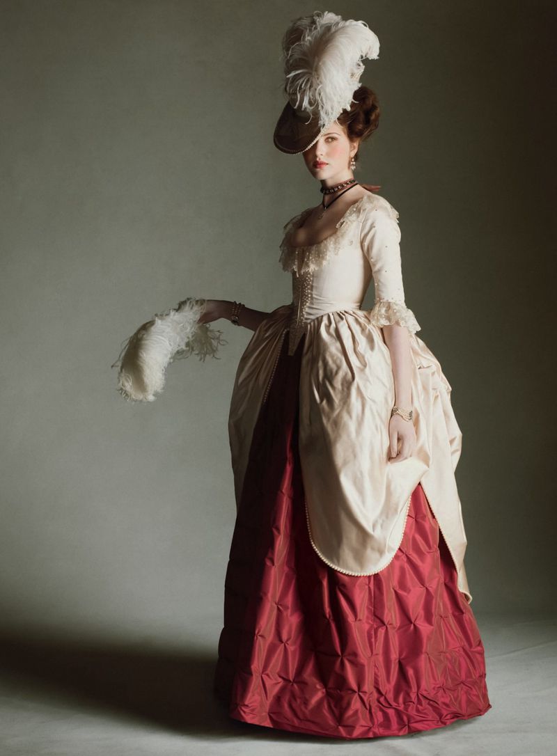 Woman in historical gown with feather hat, posing in studio, vintage style portrait, indoor setting, detailed textile and costume design.