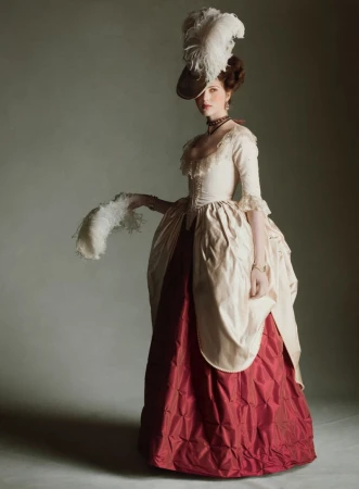 Woman in historical gown with feather hat, posing in studio, vintage style portrait, indoor setting, detailed textile and costume design.