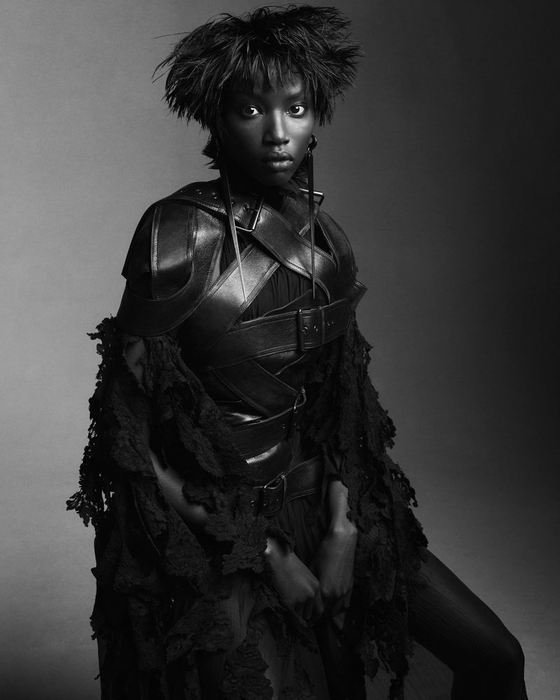 Monochrome studio portrait of a Black woman in a striking textured garment, leather-like chest piece, and feathered hair, looking at the camera.