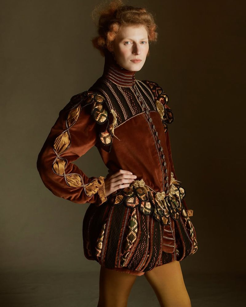 A red-haired woman poses in a Renaissance-inspired brown and gold outfit against a dark backdrop. The portrait captures the detail of the costume.