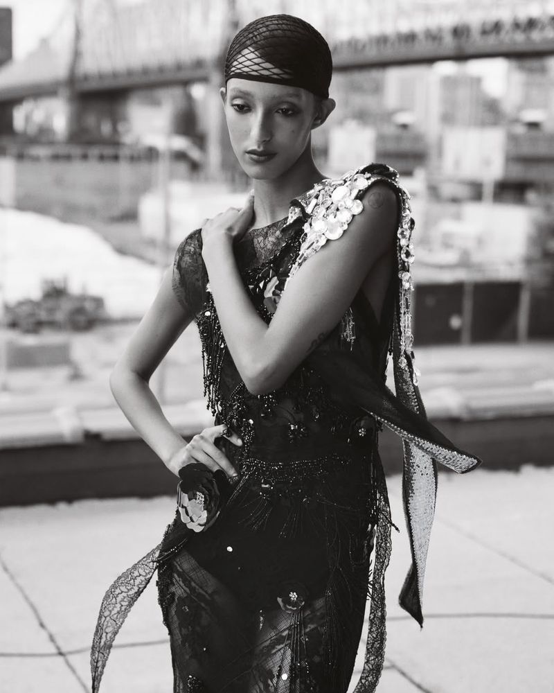 Monochrome shot of a woman in a sequined gown posing on a rooftop with a city skyline, featuring a bridge and buildings, fashion editorial.