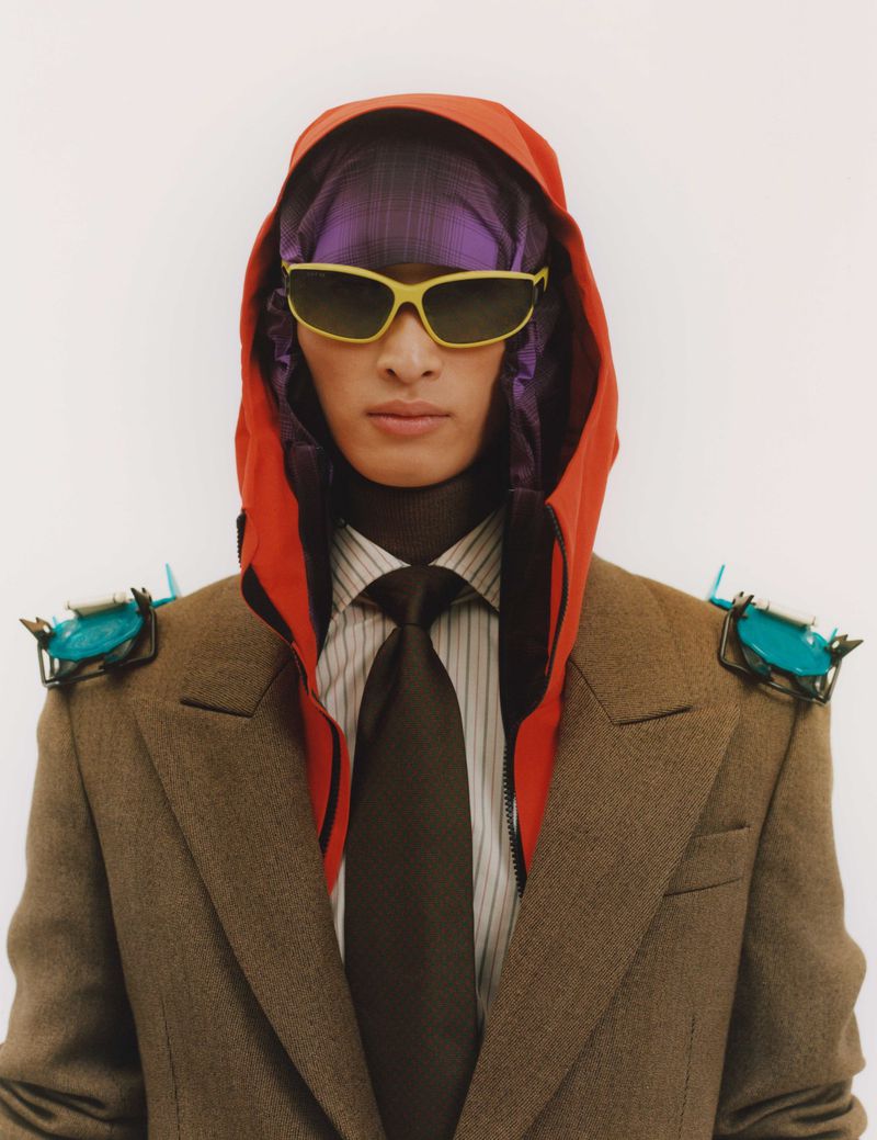 Portrait of a person wearing a brown suit, orange hood, purple head covering, and yellow sunglasses against a white background.