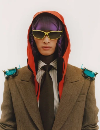 Portrait of a person wearing a brown suit, orange hood, purple head covering, and yellow sunglasses against a white background.
