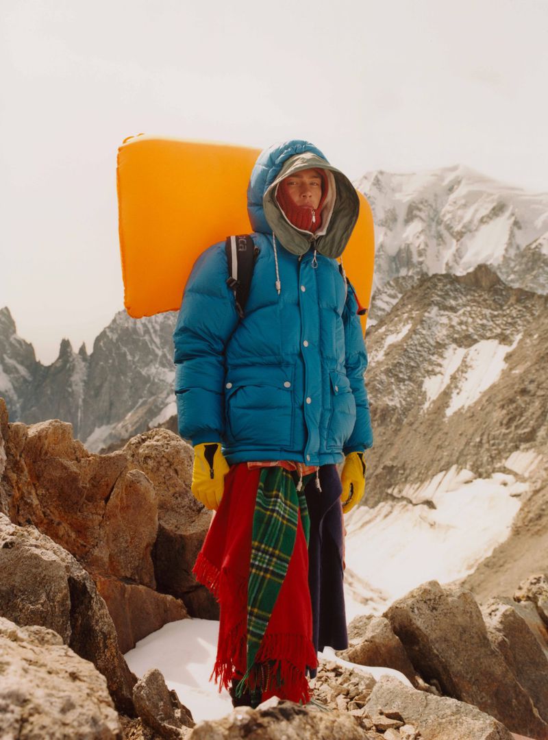 Person in a blue puffer jacket with a bright yellow inflatable pack stands on a rocky mountain with snow and distant peaks.
