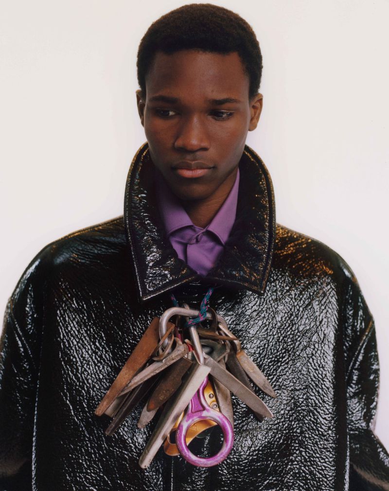 Portrait of a young adult Black male wearing a black patent leather jacket and a purple dress shirt with a cluster of keys around his neck.