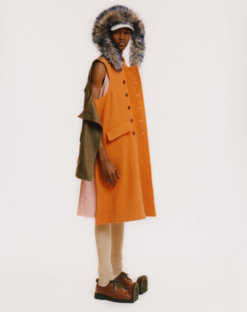 Fashionable adult in a colorful vest dress with fur-lined hood and chunky shoes poses against a stark white backdrop in a studio shot.