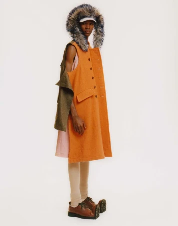 Fashionable adult in a colorful vest dress with fur-lined hood and chunky shoes poses against a stark white backdrop in a studio shot.