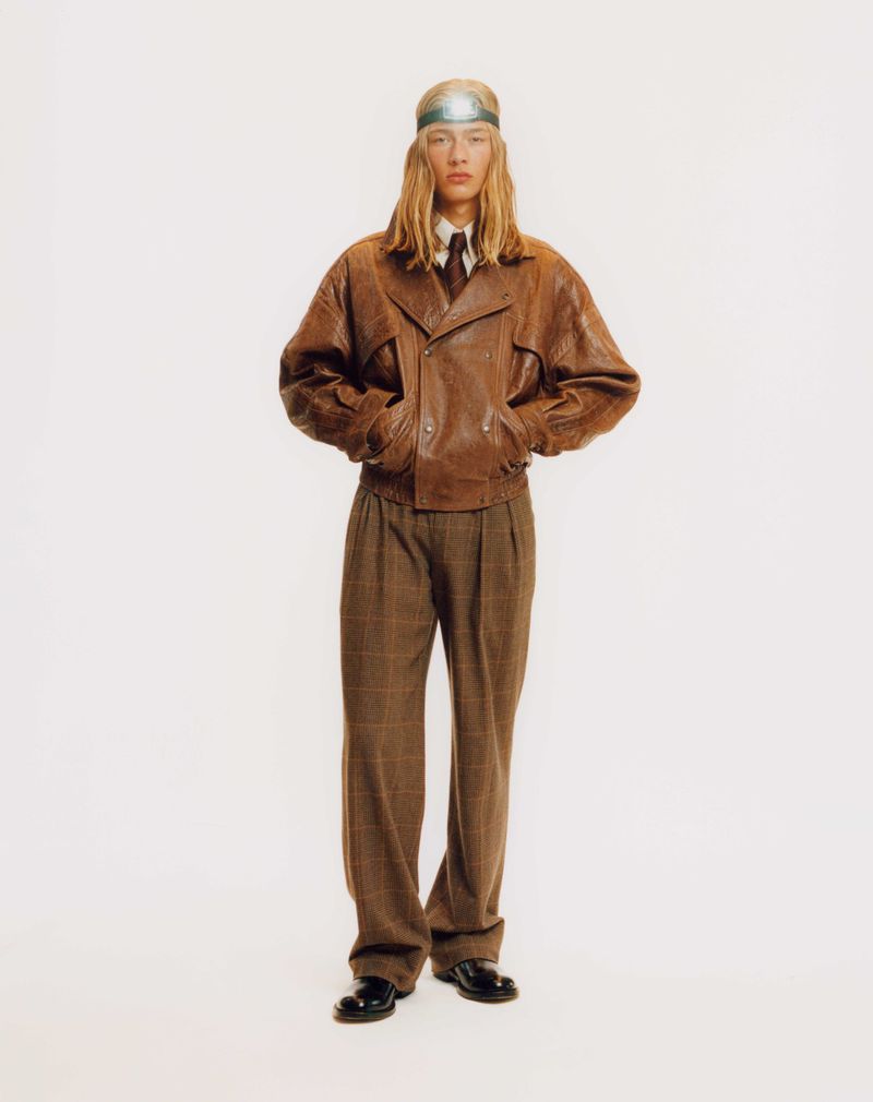 Fashionable adult male in brown leather jacket, plaid trousers, and futuristic headband, posing against a white studio backdrop.