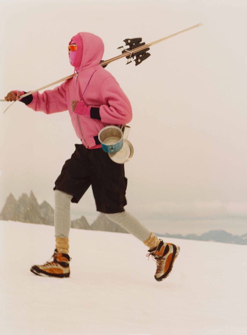 Adult in ski boots and balaclava walks across snow with skis over shoulder, carrying metal pots, with snowy mountains behind.
