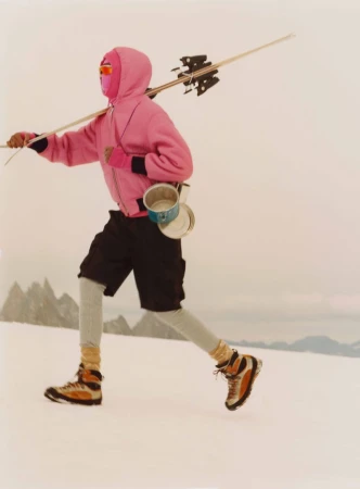 Adult in ski boots and balaclava walks across snow with skis over shoulder, carrying metal pots, with snowy mountains behind.