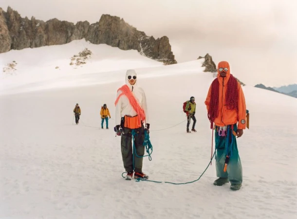 Group of six mountain climbers traverses a snowy mountain with peaks in the background, wearing orange, white, and yellow jackets.