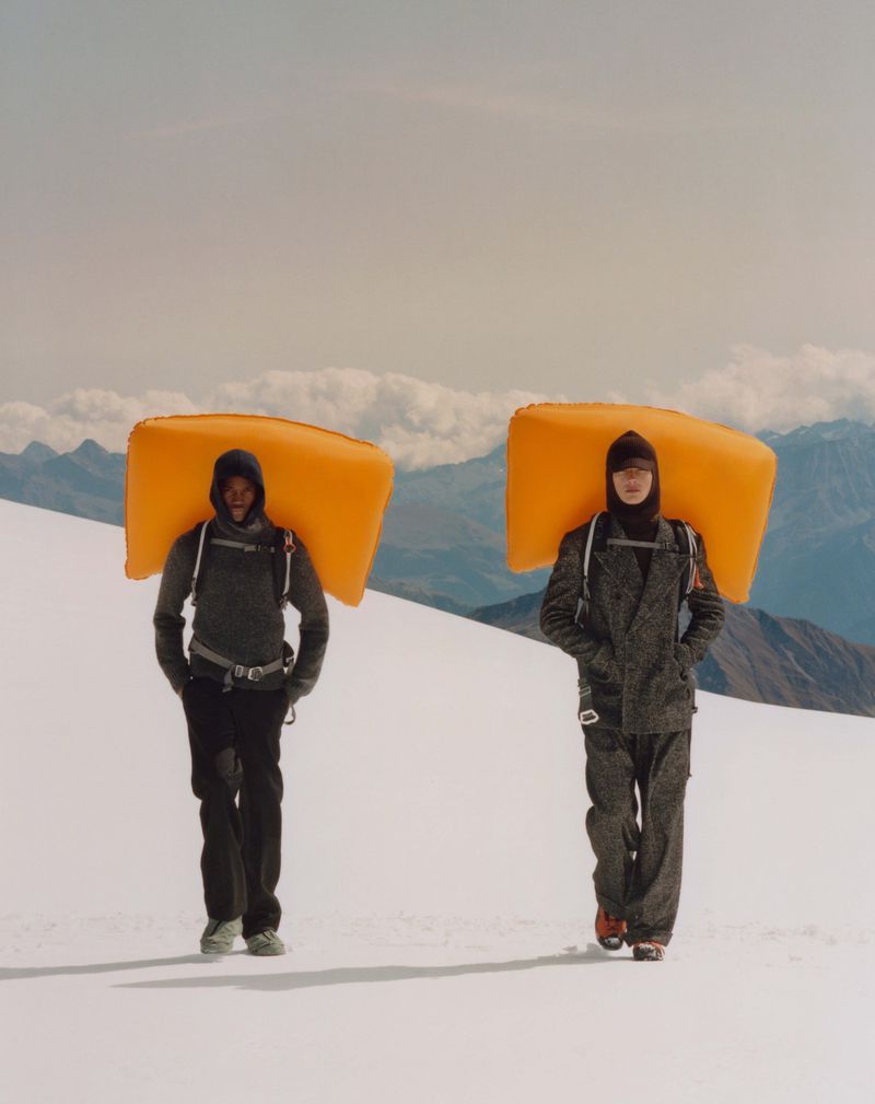 Two people walk through a snow-covered mountain landscape with orange pads on their backs, wearing grey and black jackets and pants.