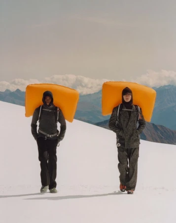 Two people walk through a snow-covered mountain landscape with orange pads on their backs, wearing grey and black jackets and pants.