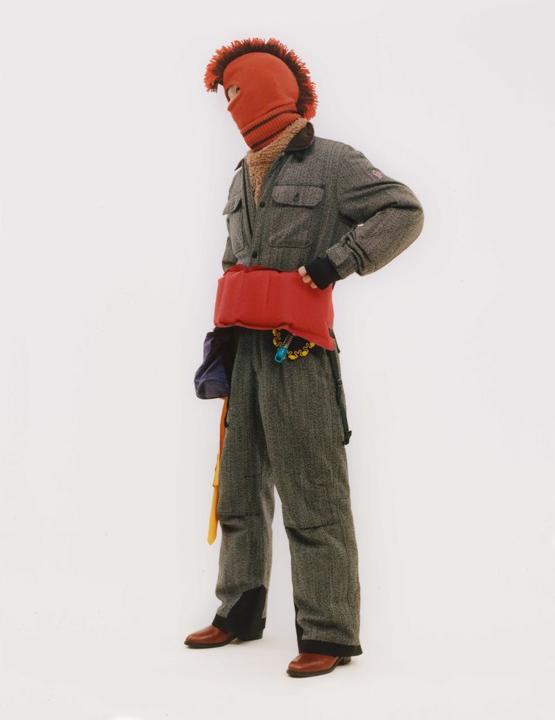 Person stands in gray herringbone jumpsuit, red balaclava with orange trim, and red quilted hip pack against a stark white backdrop.