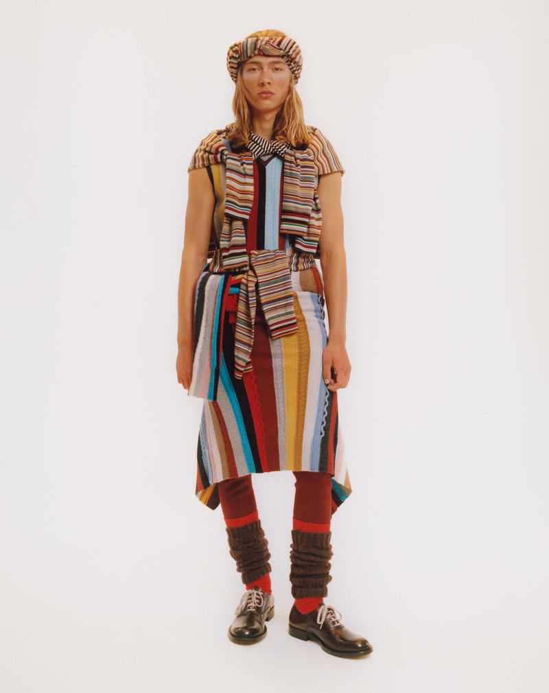 Full-shot of a person wearing a multicolored striped outfit with matching hat and leg warmers, posing against a white background.