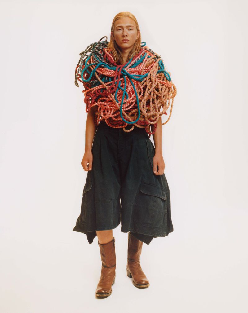 Full shot of a blonde person in black culottes and brown boots. Their top is covered by a mass of colorful wound rope.