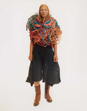 Full shot of a blonde person in black culottes and brown boots. Their top is covered by a mass of colorful wound rope.