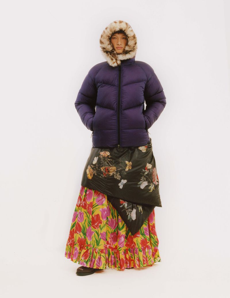 A person stands against a white background wearing a purple puffer jacket with fur hood, a black floral wrap, and a colorful floral skirt.