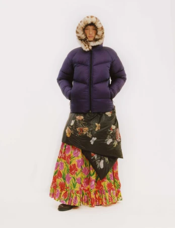 A person stands against a white background wearing a purple puffer jacket with fur hood, a black floral wrap, and a colorful floral skirt.