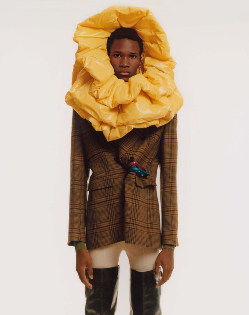 Fashionable black male model in brown plaid blazer, light pants, black boots, and a large yellow floral neckpiece posing indoors.